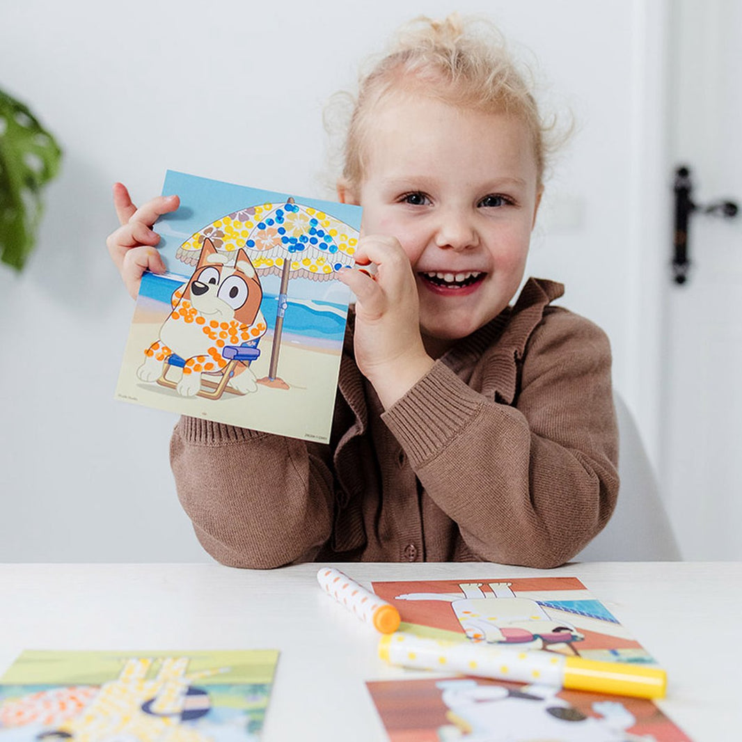 Дете с усмивка държи рисунка от творчески комплект - оцветяване с точки Bluey.